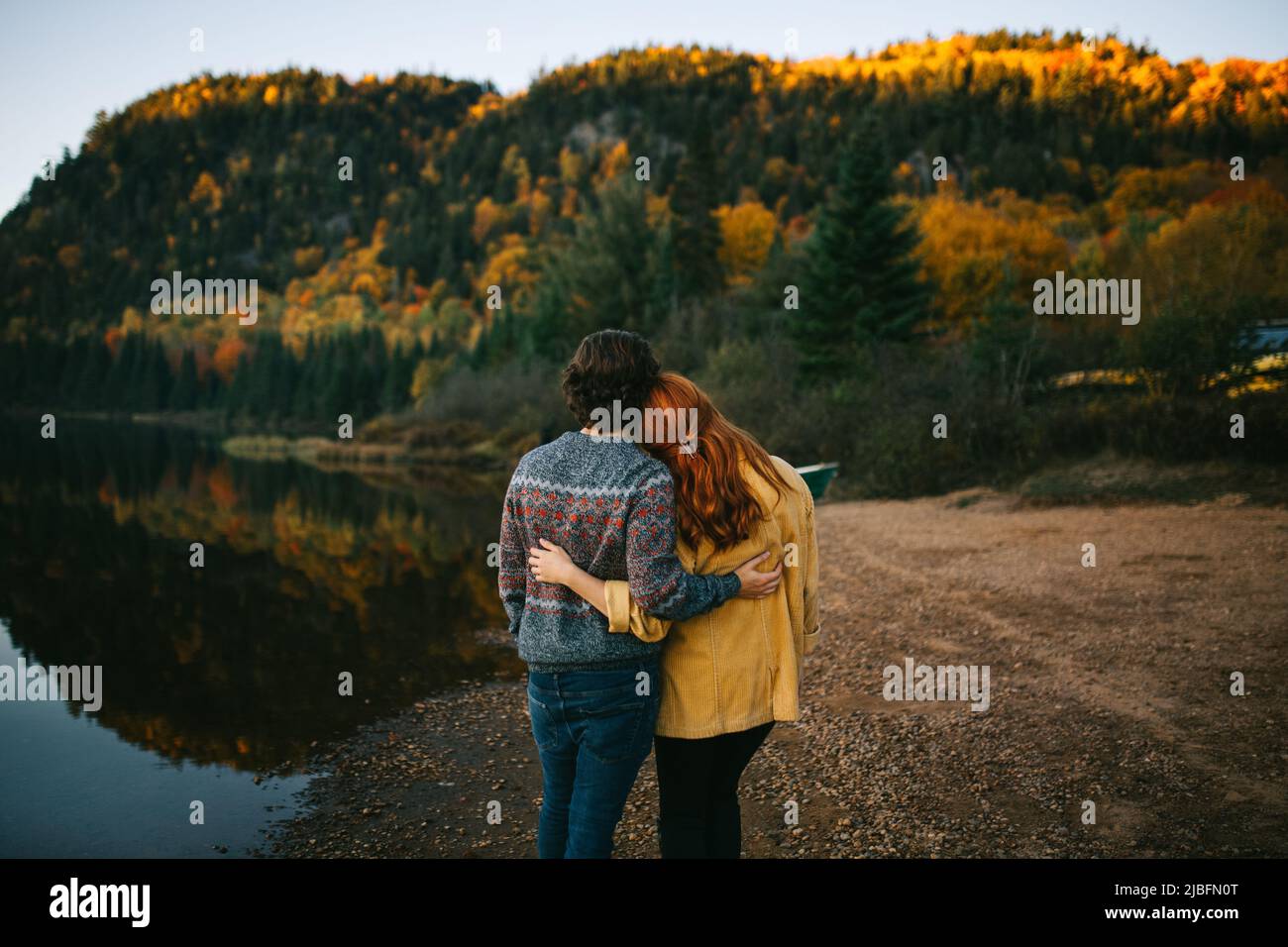 Back view of romantic unrecognizable traveling couple in stylish clothes embracing and admiring scenic autumn landscape of calm lake surrounded by lus Stock Photo