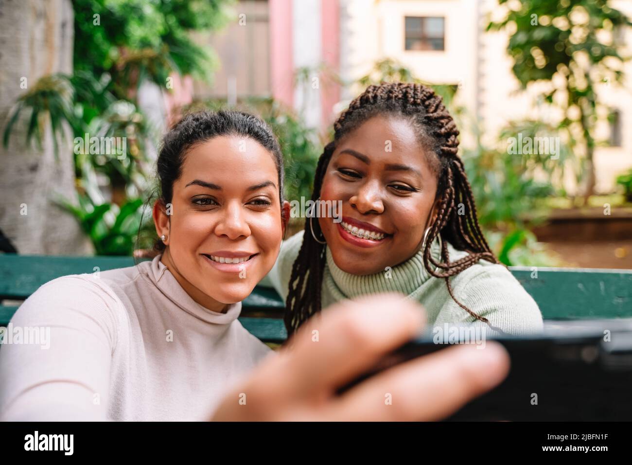 Cheerful black woman in sweater laughing and taking selfie with Hispanic girlfriend while sitting on bench in park Stock Photo