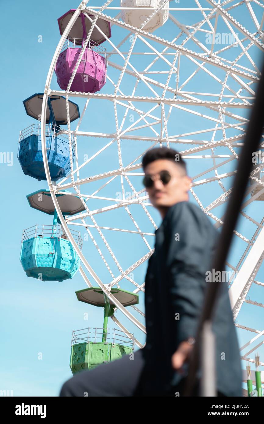 Low angle side view of male manager in smart casual clothes sitting on railing against blue sky and colorful Ferris wheel on sunny day in amusement pa Stock Photo