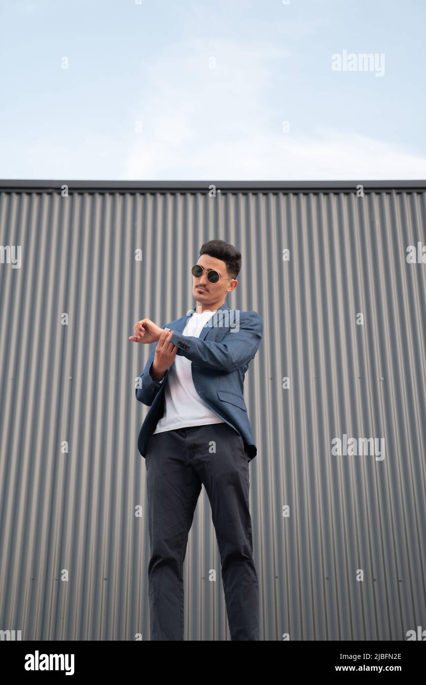 From below ethnic male entrepreneur in suit and sunglasses looking at wristwatch and checking time while standing near metal wall and waiting for meet Stock Photo