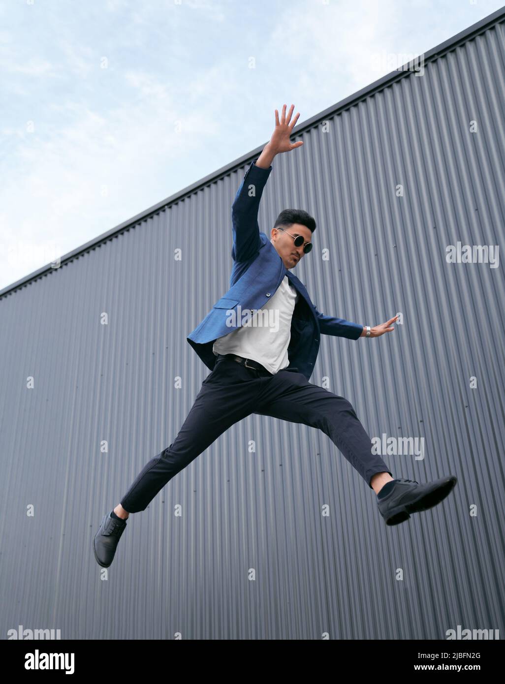 From below ethnic businessman in suit and sunglasses spreading arms and leaping against metal wall on sunny day on city street Stock Photo