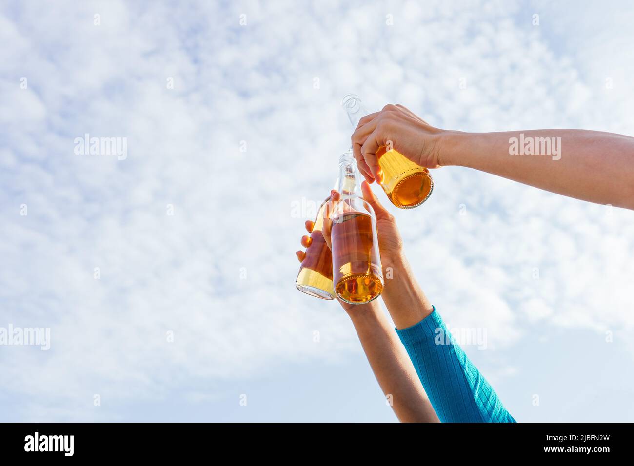 From below anonymous cropped hands of man and women raising bottles of beer while chilling on beach against blue sky on summer weekend day Stock Photo