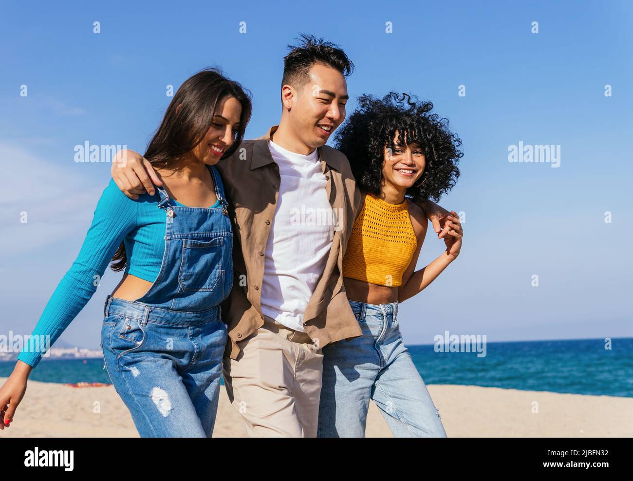 Happy Asian man embracing diverse girlfriends and laughing while walking on sandy beach against blue sky on summer weekend day on resort Stock Photo