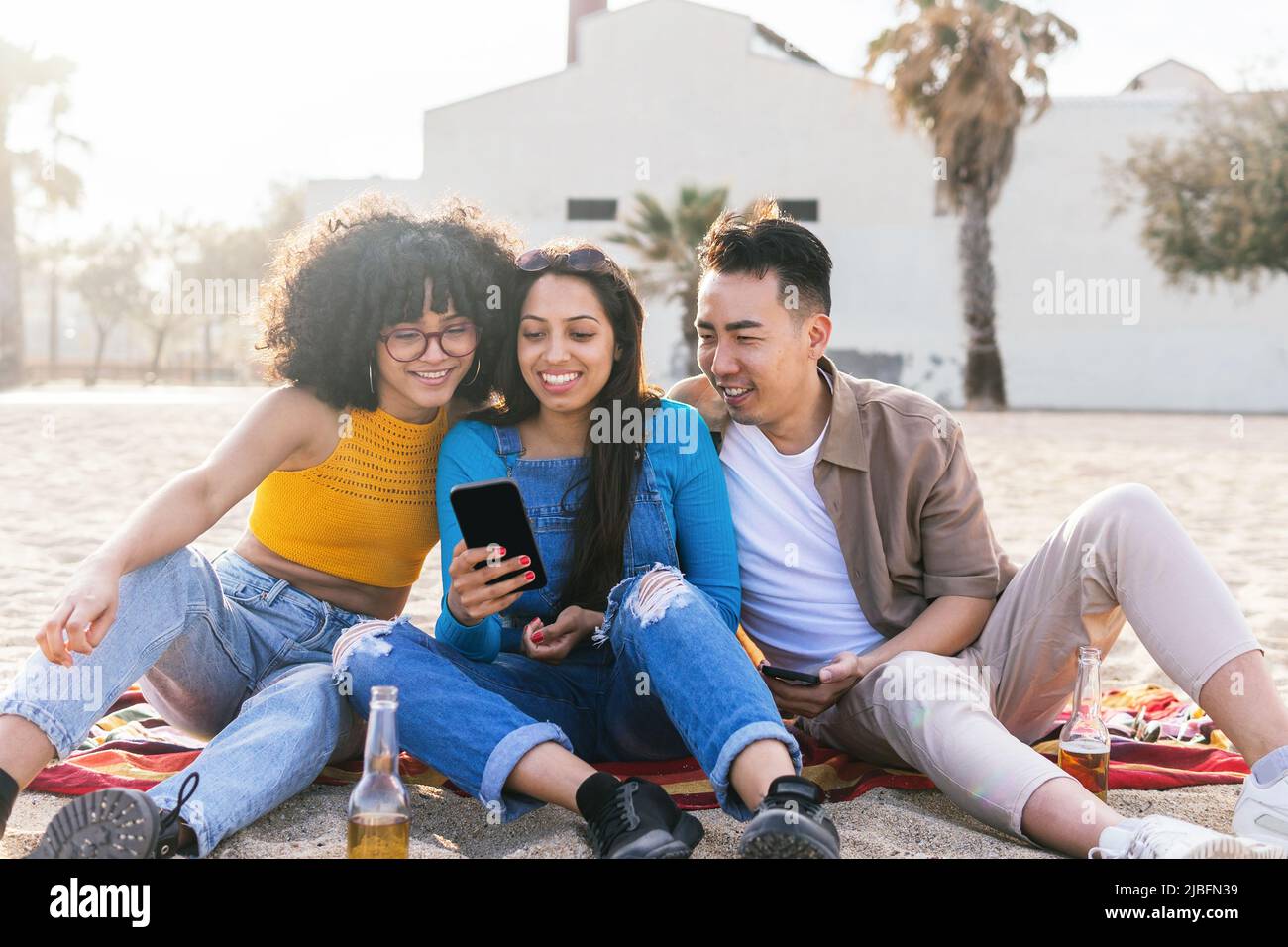 Happy multiracial man and women smiling and browsing social media on cellphones while resting on beach on sunny summer day on resort Stock Photo