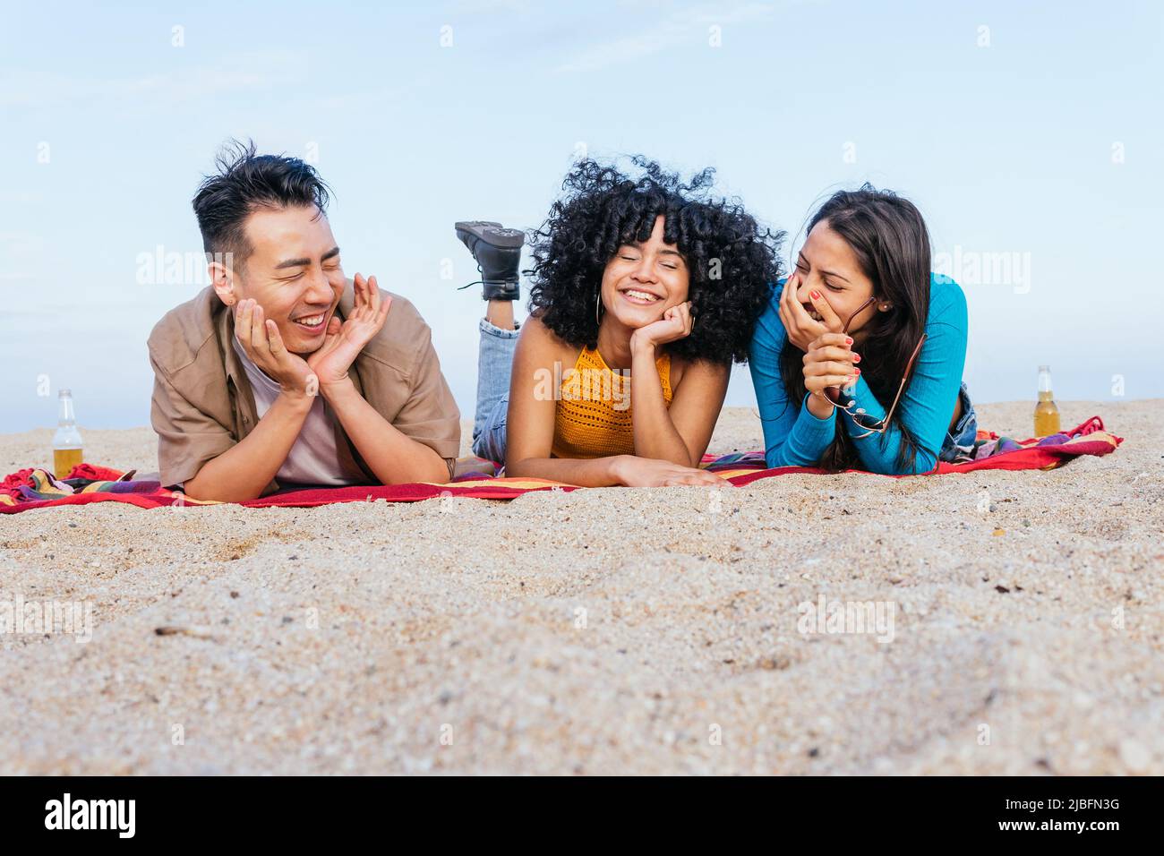 Diverse man and women in casual clothes lying on blanket with eyes closed and smiling while spending time on sandy beach near sea on summer weekend da Stock Photo