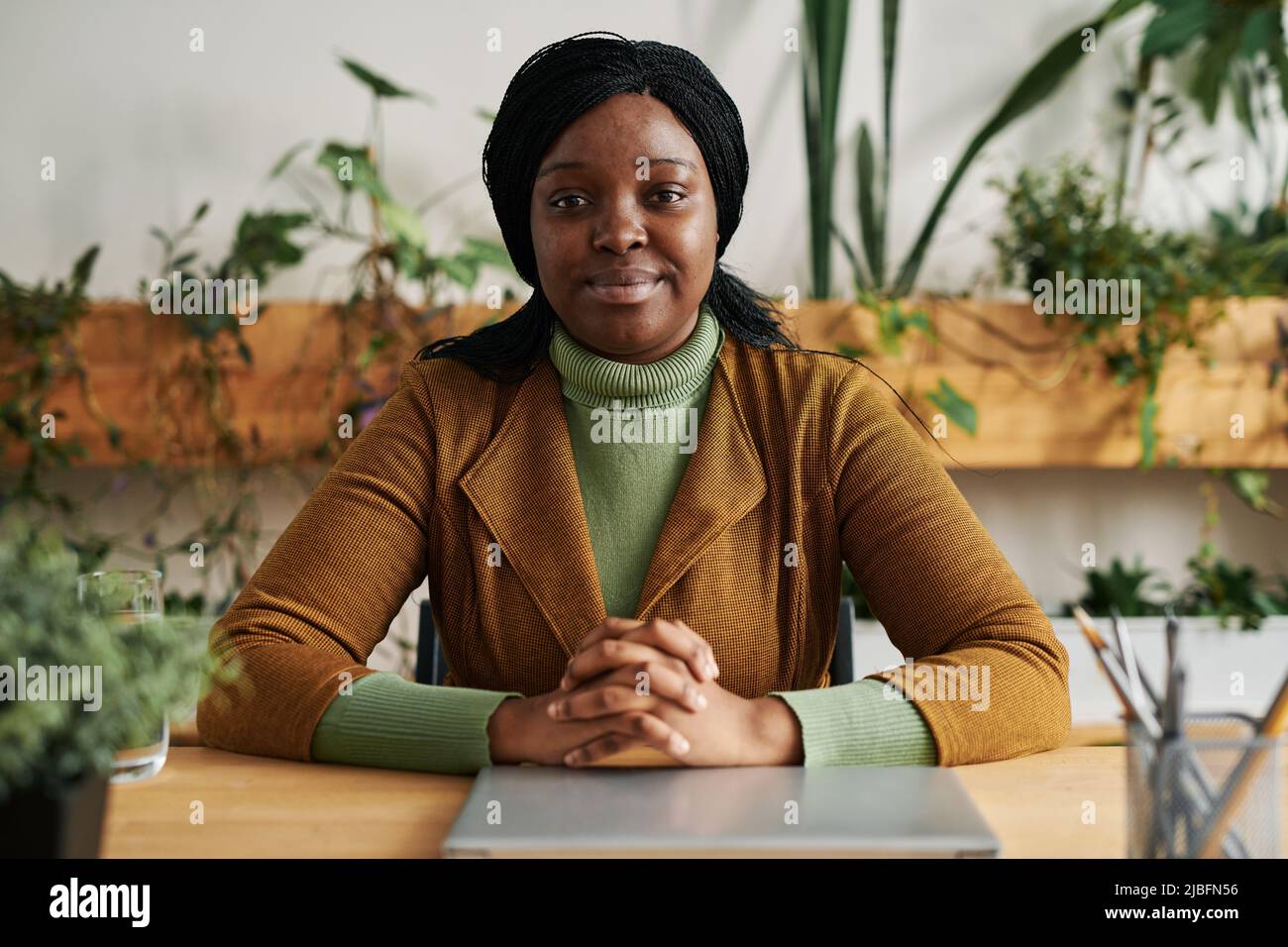 Young confident African American female psychologist in smart casualwear sitting by workplace in office and looking at camera Stock Photo