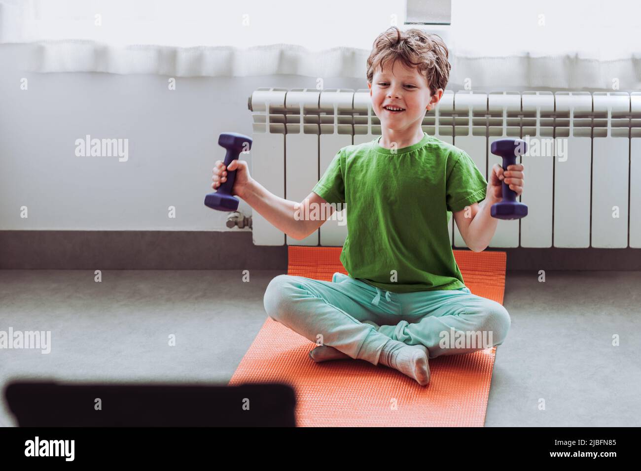 Merry child in activewear smiling and doing exercise with dumbbells while sitting cross legged on mat and watching tutorial video on tablet during onl Stock Photo