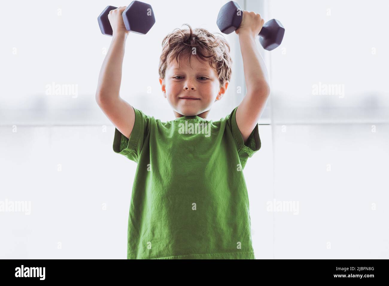 Happy kid in activewear smiling and exercising with dumbbells against window during fitness training in morning at home Stock Photo