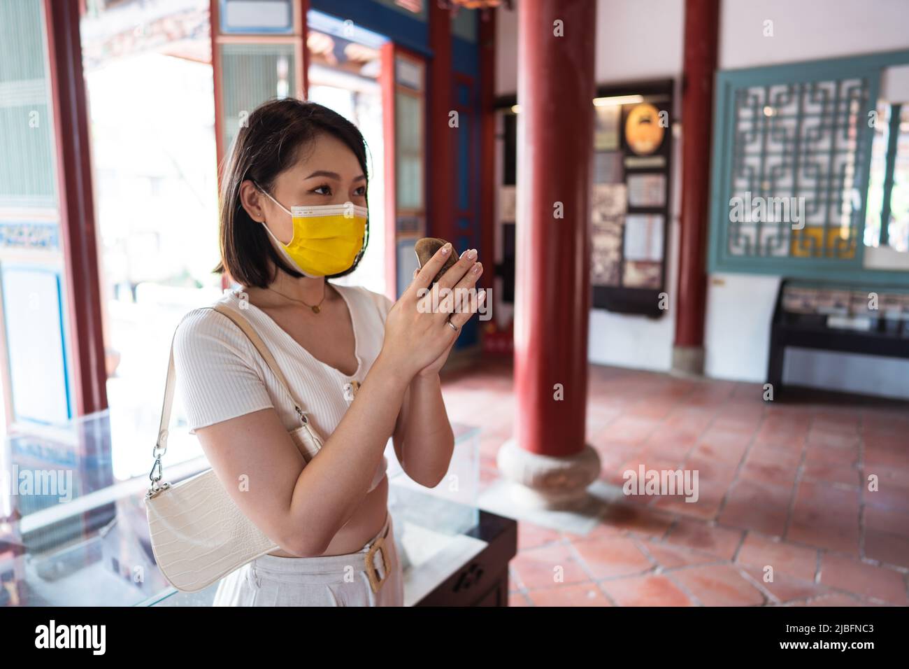 Concentrated young Asian female traveler with dark hair in crop top and face mask holding moon blocks in hands and looking away while standing in temp Stock Photo