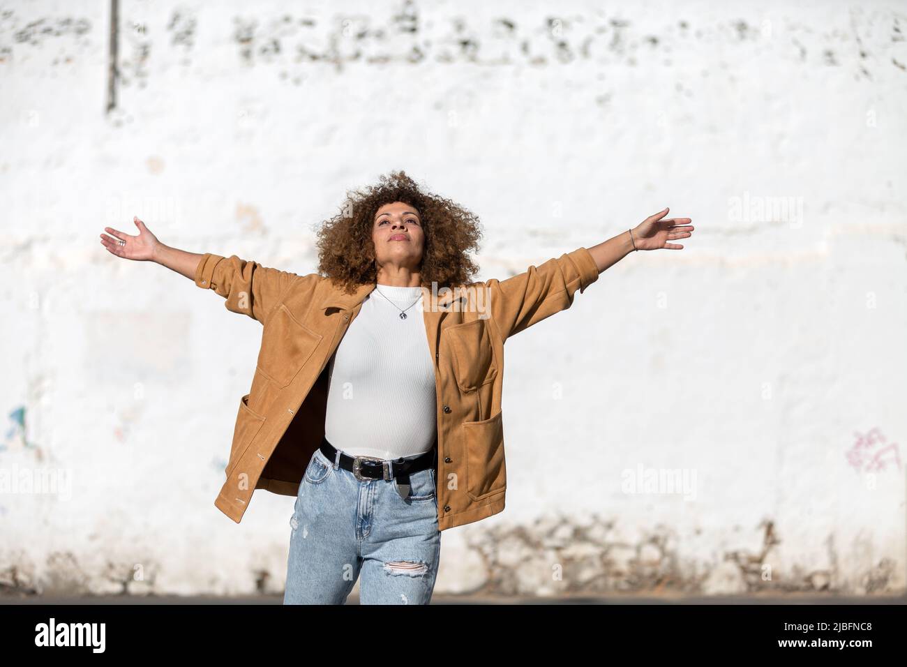 Adult Hispanic female in casual clothes with curly hair stretching out arms and inhaling air against grungy wall on sunny day on city street Stock Photo