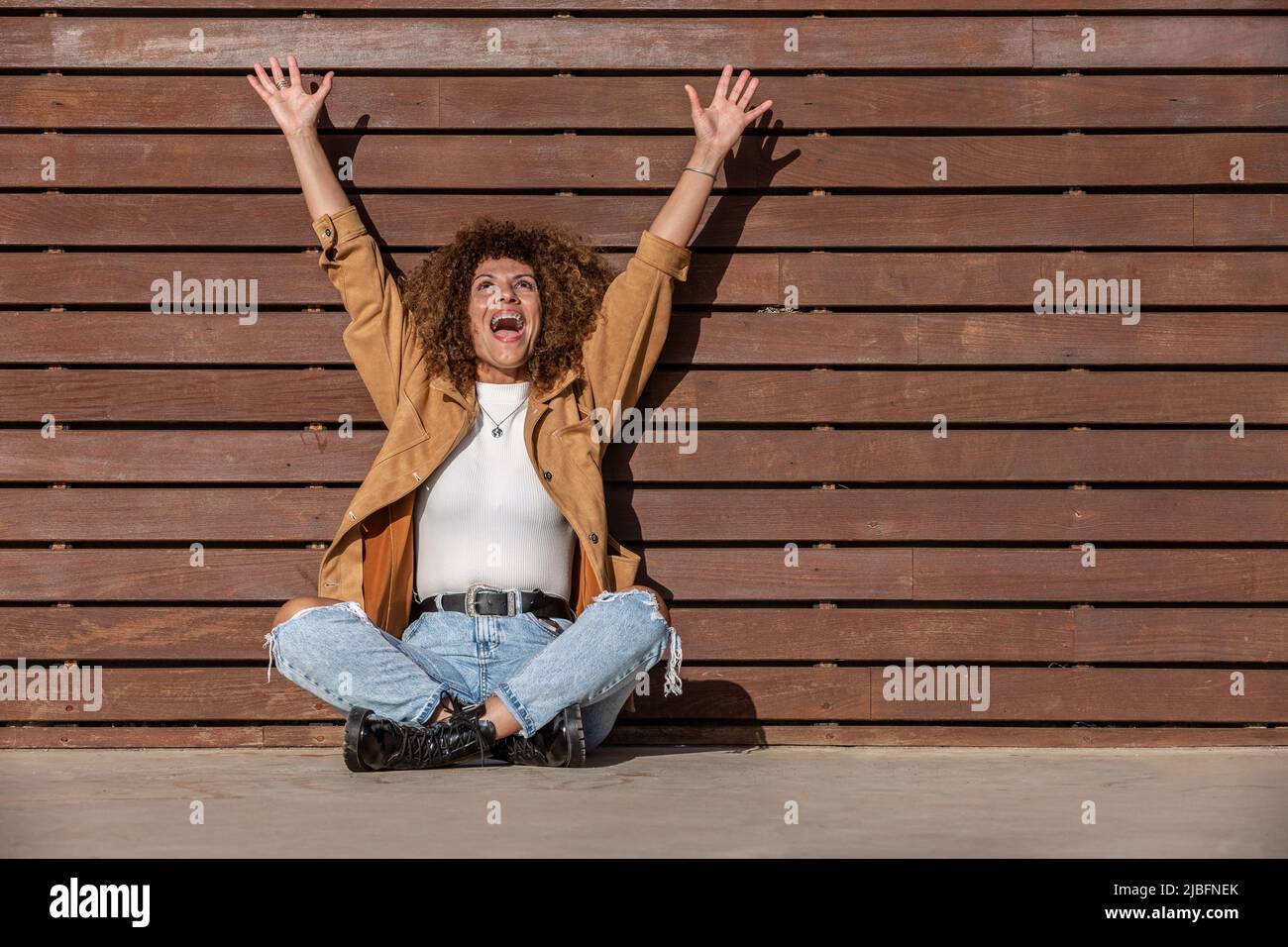 Cheerful adult Hispanic woman in casual clothes raising arms and screaming loudly while sitting cross legged on ground against brown wooden wall on su Stock Photo