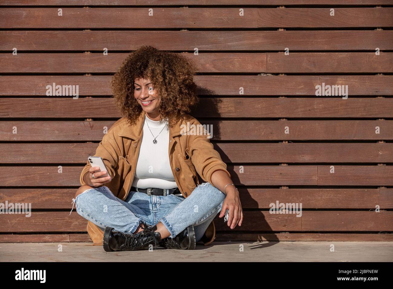 Positive adult Hispanic female in casual clothes with curly hair smiling and browsing social media on cellphone while sitting cross legged on ground n Stock Photo