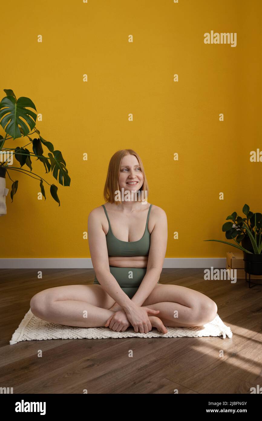 Full body of confident positive young lady in underwear with red hair smiling and looking away while sitting on floor in Lotus pose during yoga practi Stock Photo