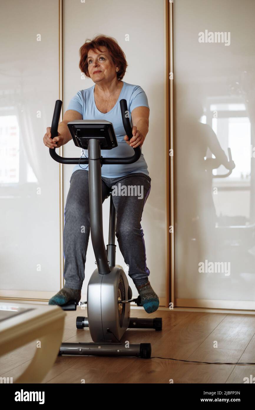 Portrait of elderly woman wearing blue T-shirt, grey trousers, sitting on stationary bicycle, working out exercises. Stock Photo