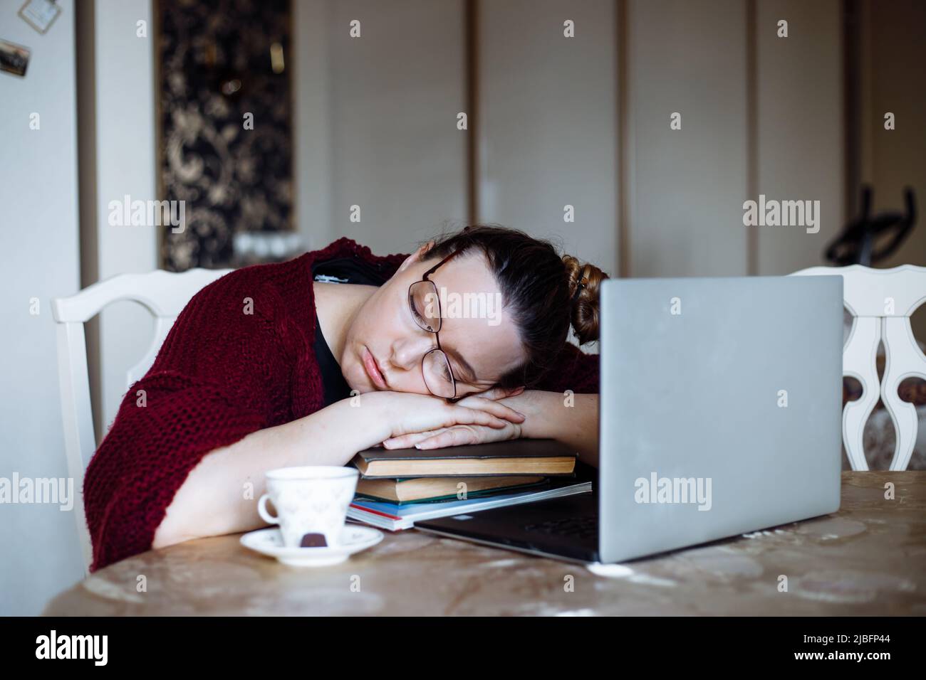Portrait of tired middle-aged woman sitting at table near laptop, sleeping on crossed arms on pile of book sat home.  Stock Photo