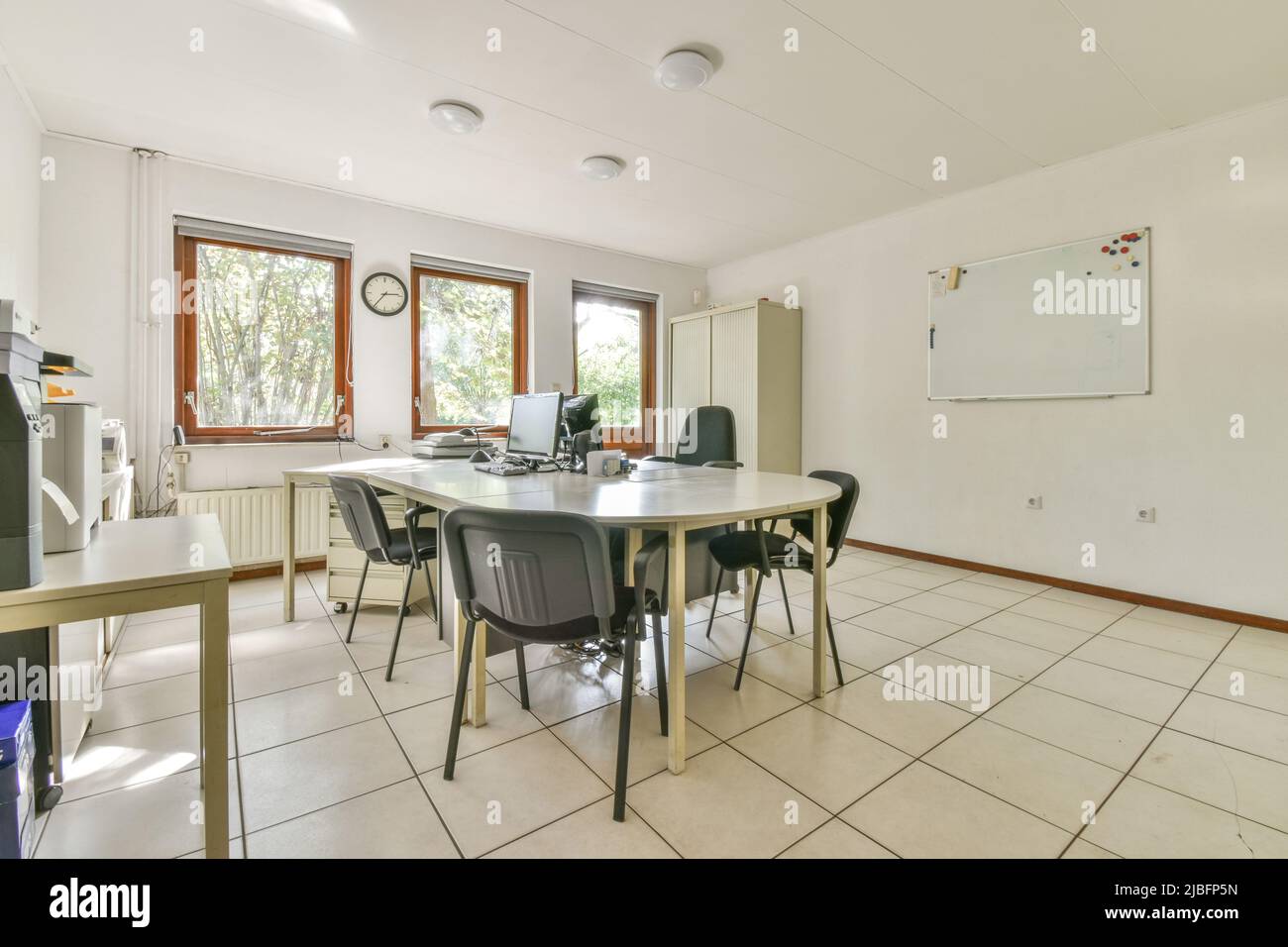 Chairs at oval table with modern computers in spacious office with whiteboard hanging on wall and tiled floor on sunny day Stock Photo