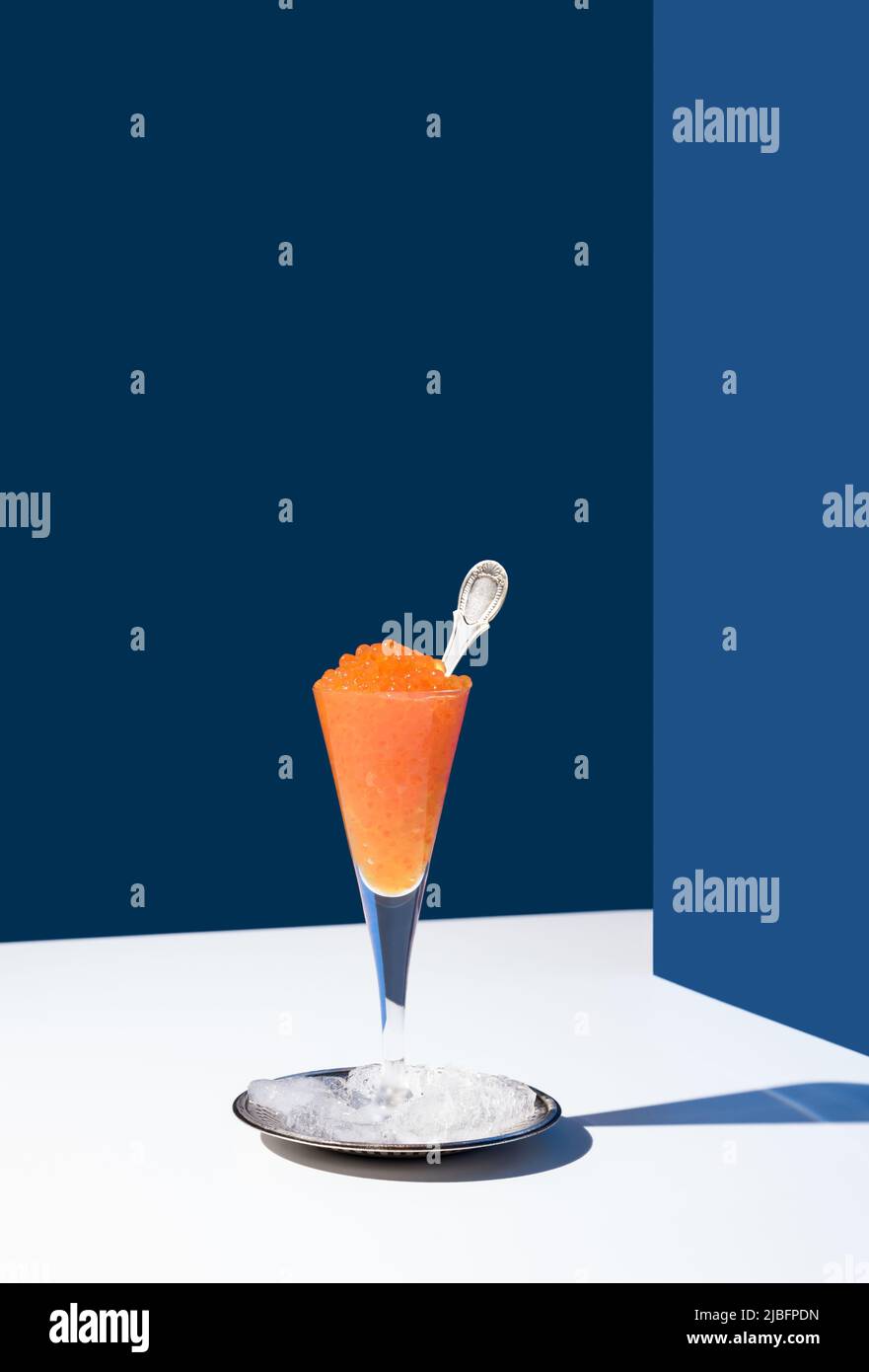 Orange tobiko caviar served on cocktail glass placed on silver plate on ice with spoon on white and blue background Stock Photo