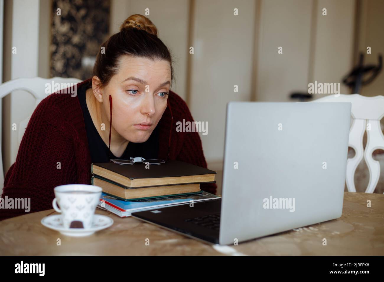 Concentrated tired woman reading info, drink coffee, using laptop, remote job at home. Work problems at freelance office Stock Photo