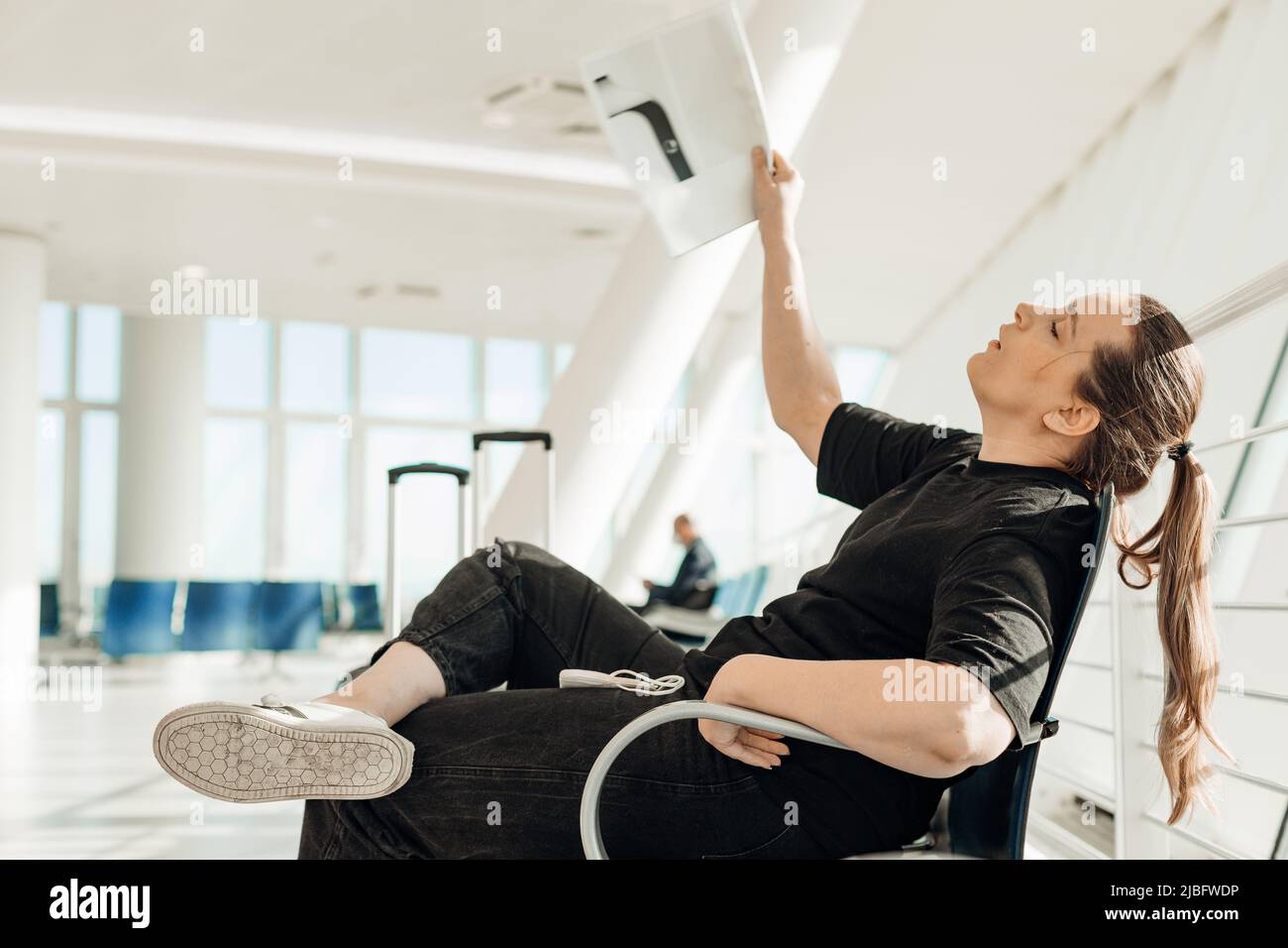 Side view of woman sitting on chair in transit lounge of airport terminal, waving with magazine, suffering from heat. Stock Photo