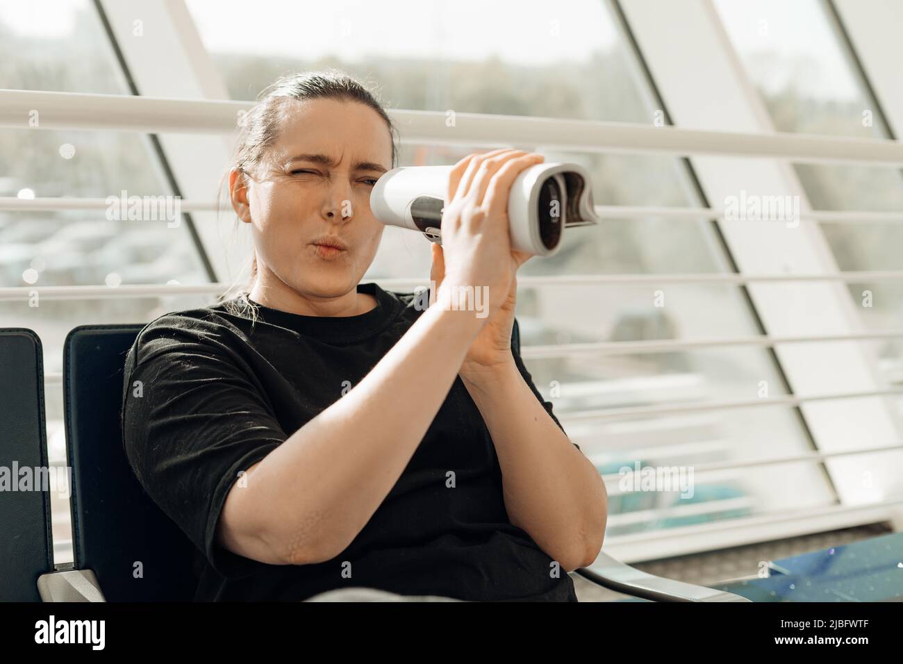 Portrait of woman passenger sitting in transit lounge looking through telescope made of rolled-up magazine, squinting. Stock Photo