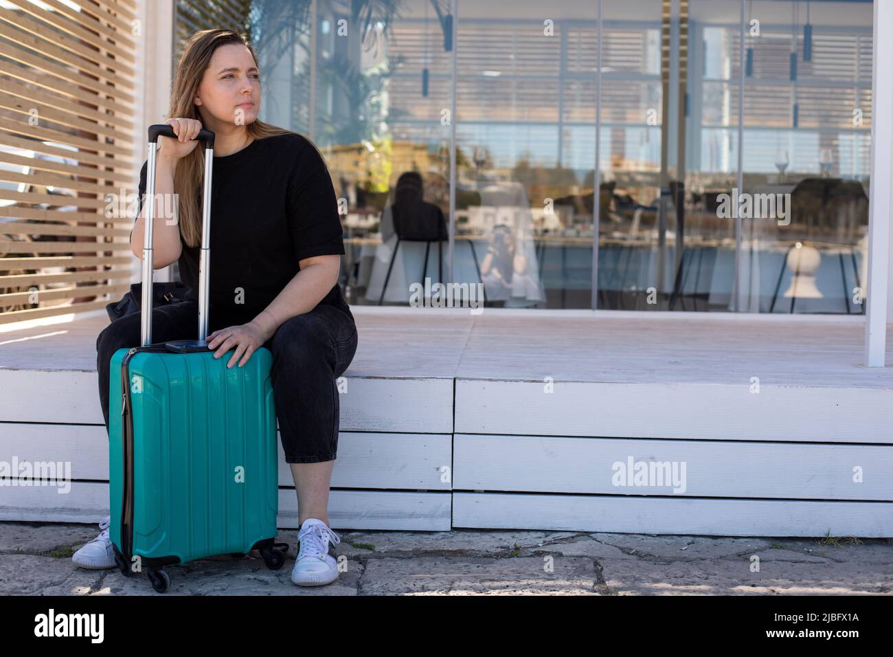 Tired blond woman in casual clothes with luggage, bags, sitting near airport terminal, outside wait room. Late arrival Stock Photo