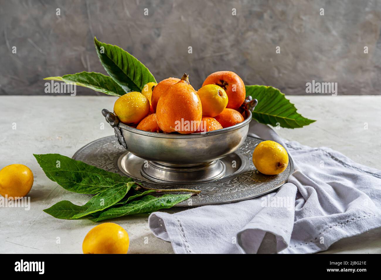 Medlar fruits on antique tin bowl and plate with leaves over concrete background, grey napkin.  Stock Photo