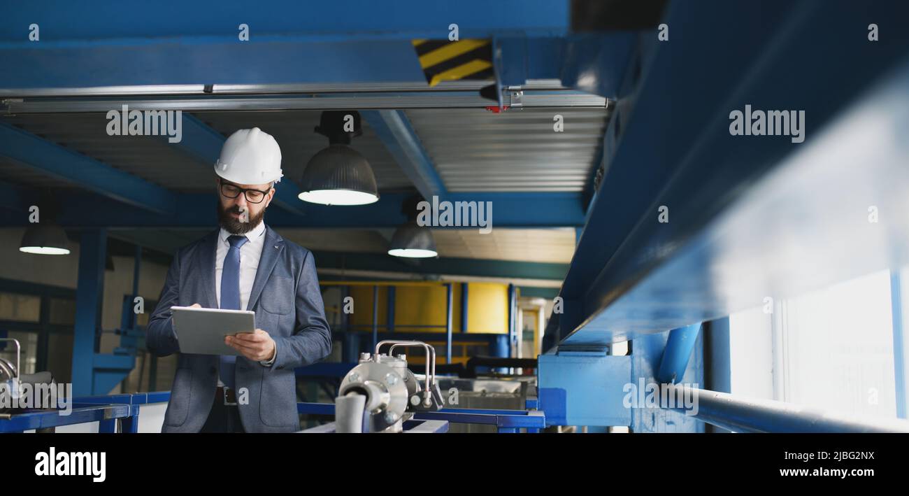 Chief Engineer in the hard hat walks through industrial factory while holding tablet. Stock Photo