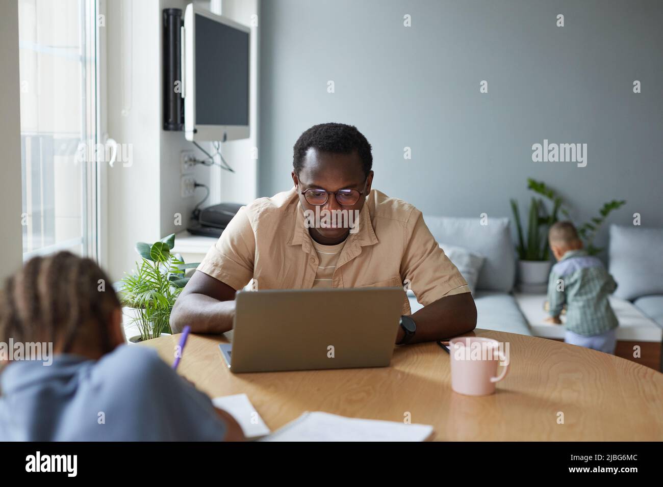 Portrait of black stay at home father using laptop with two kids in background, copy space Stock Photo