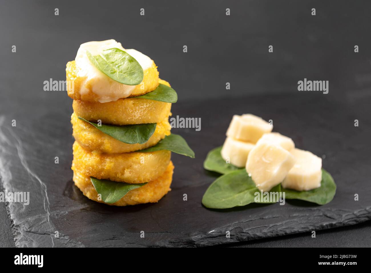 Italian porridge fried in the form of polenta pancakes on a black background with macarella cheese and spinach Stock Photo