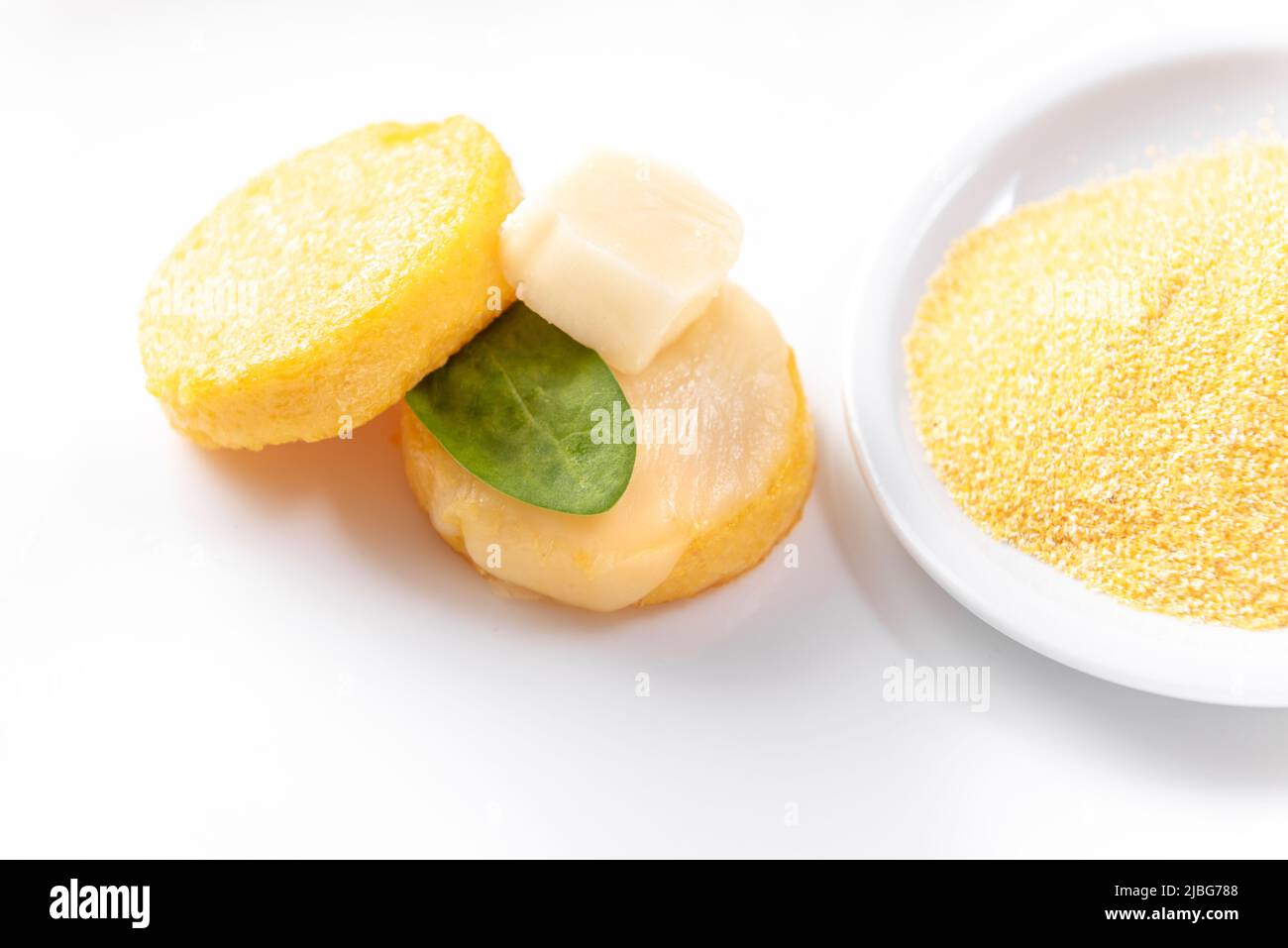 fried polenta in the form of pancakes on a white background with cheese and spinach next to a plate of raw corn grits Stock Photo