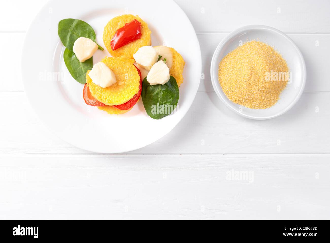 fried polenta in the form of pancakes on a white background with cheese and spinach next to a plate of raw corn grits Stock Photo