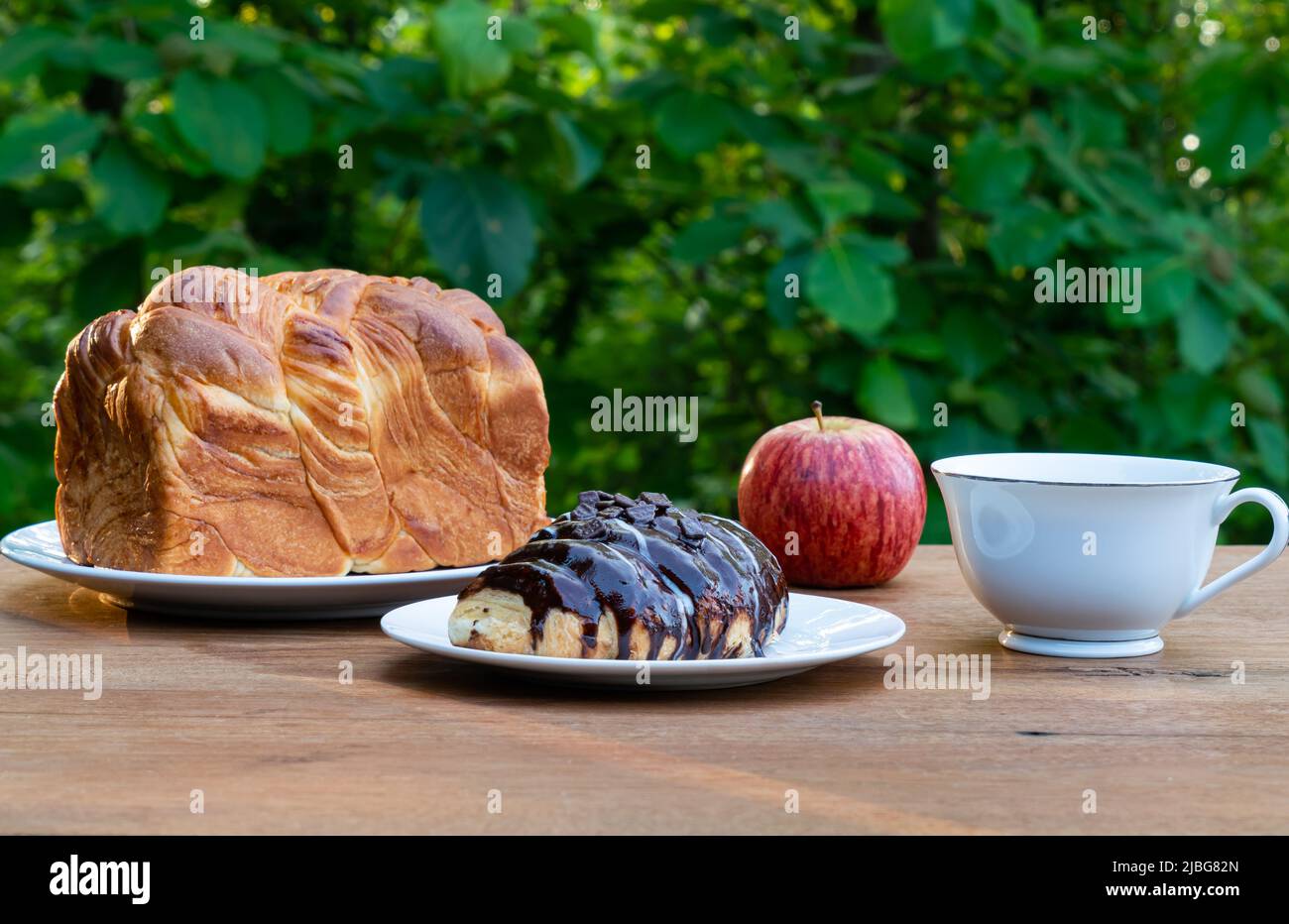 Breakfast at a green teak forest background. Breakfast with a loaf of homemade bread in white ceramic dish, a chocolate croissant, a ripe red apple an Stock Photo