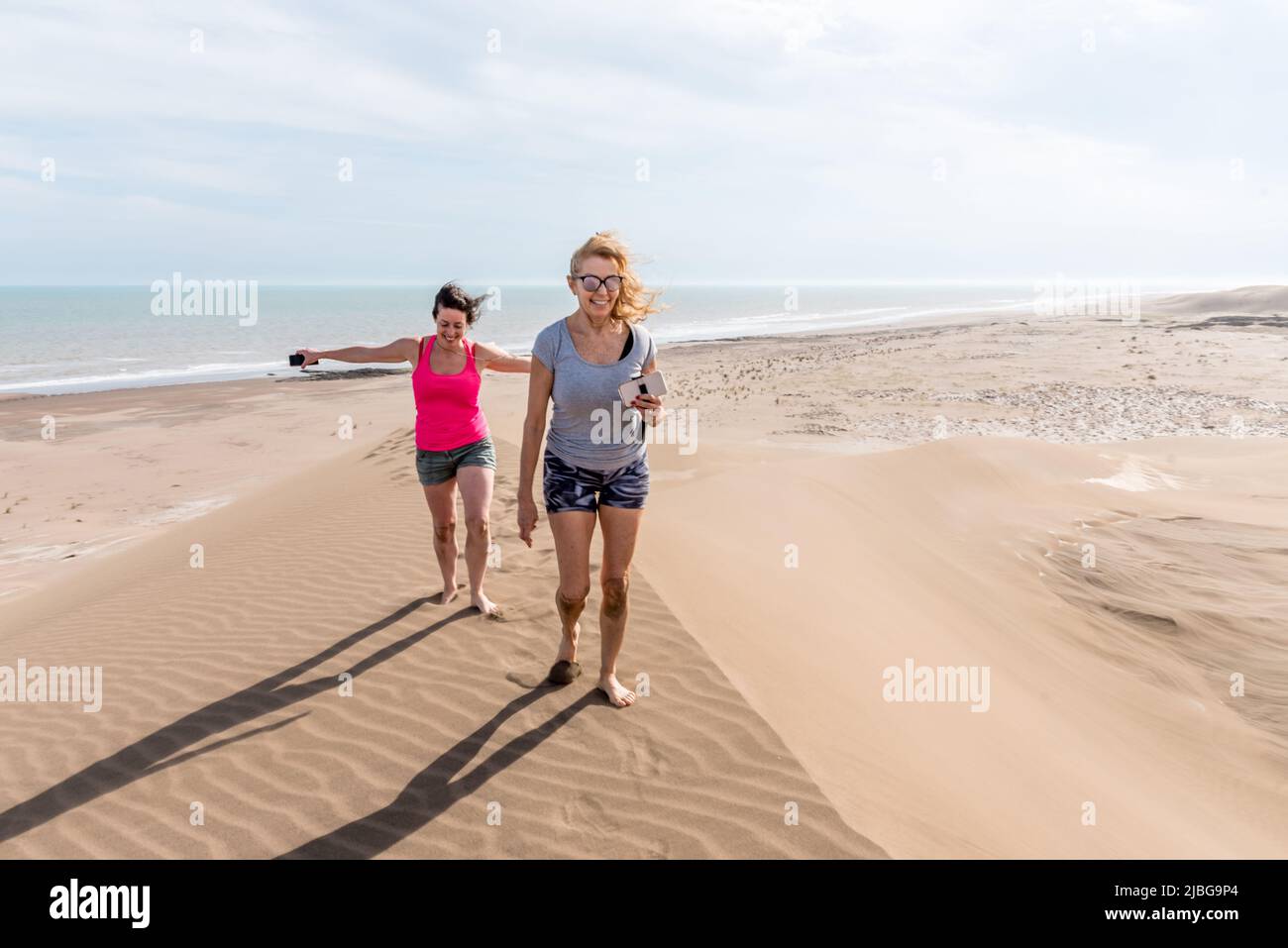 Front view of two women walking on top of a large dune, one of them with arms open to the wind. Mother and daughter Stock Photo
