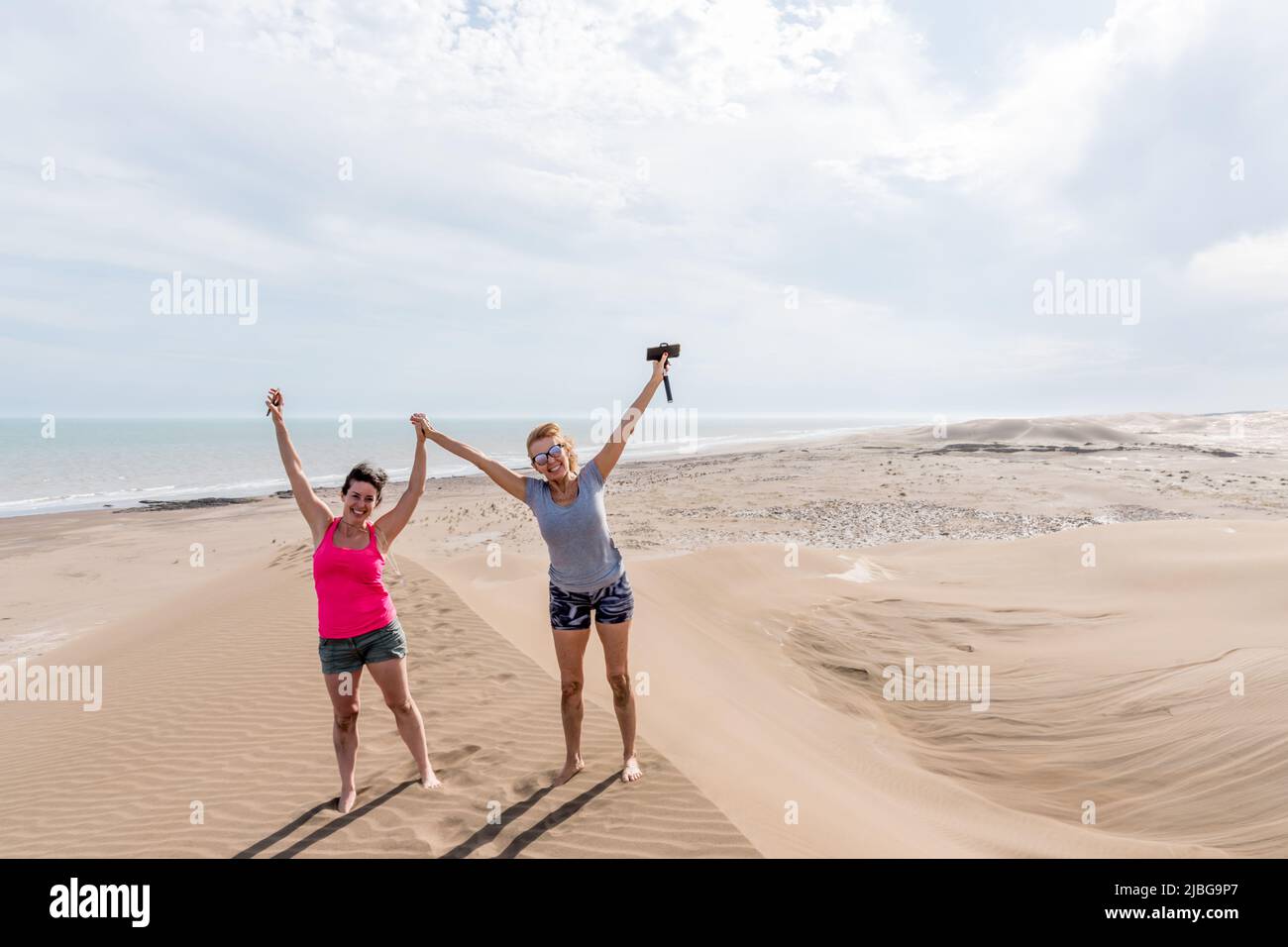 Mature mother and adult daughter with their arms raised, on a large dune with the beach in the background. Stock Photo