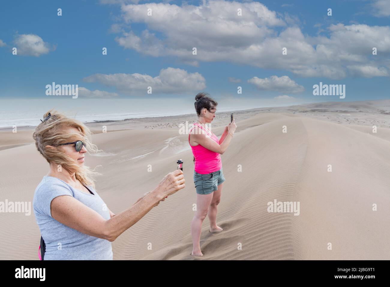Side view of mature mother and adult daughter taking pictures on top of a big dune with the sea and the beach behind Stock Photo