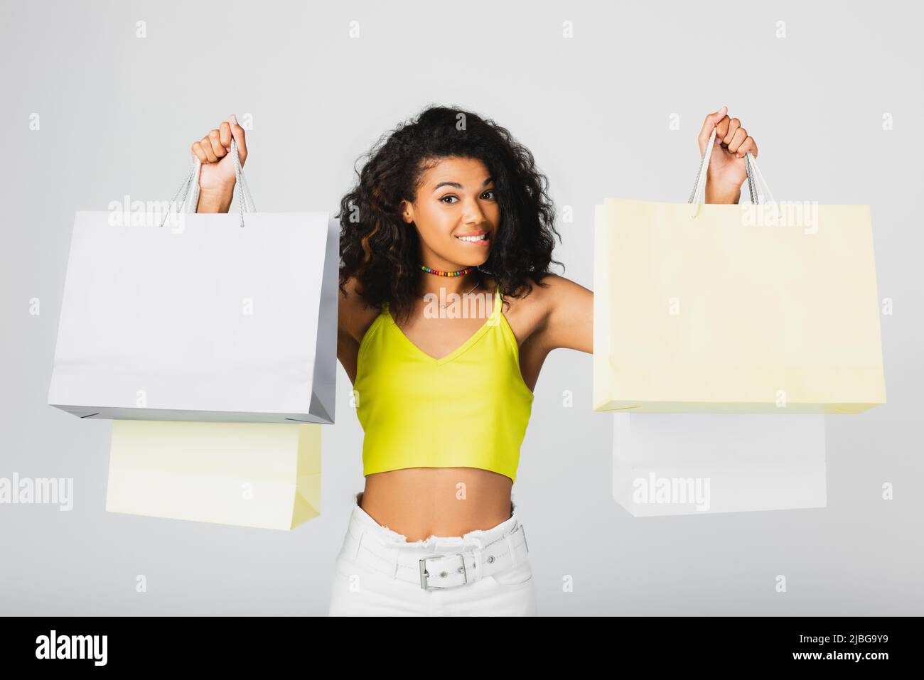 cheerful african american woman holding shopping bags and biting lip isolated on grey Stock Photo