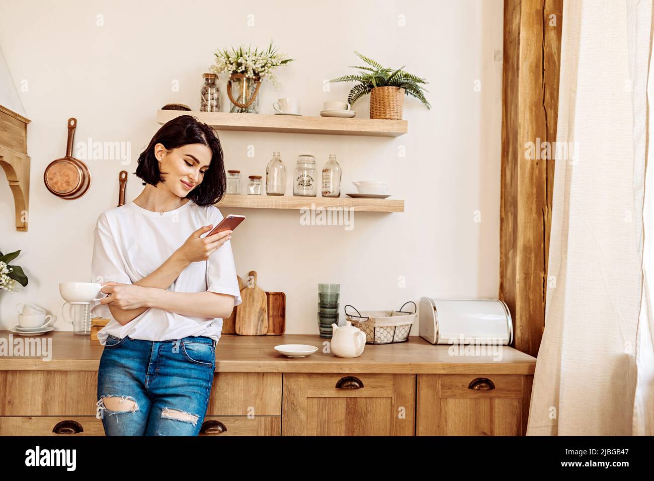 Lifestyle portrait of a happy young girl browsing the news on the phone over a cup of tea or coffee in the kitchen early in the morning Stock Photo