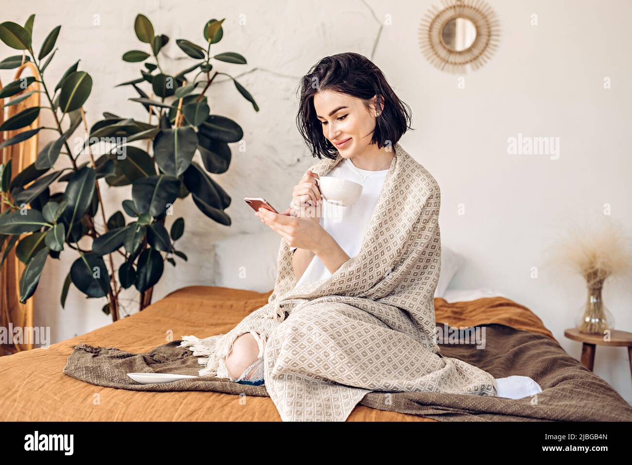 Lifestyle portrait of a happy young girl browsing the news on the phone over a cup of tea or coffee in bed early in the morning Stock Photo