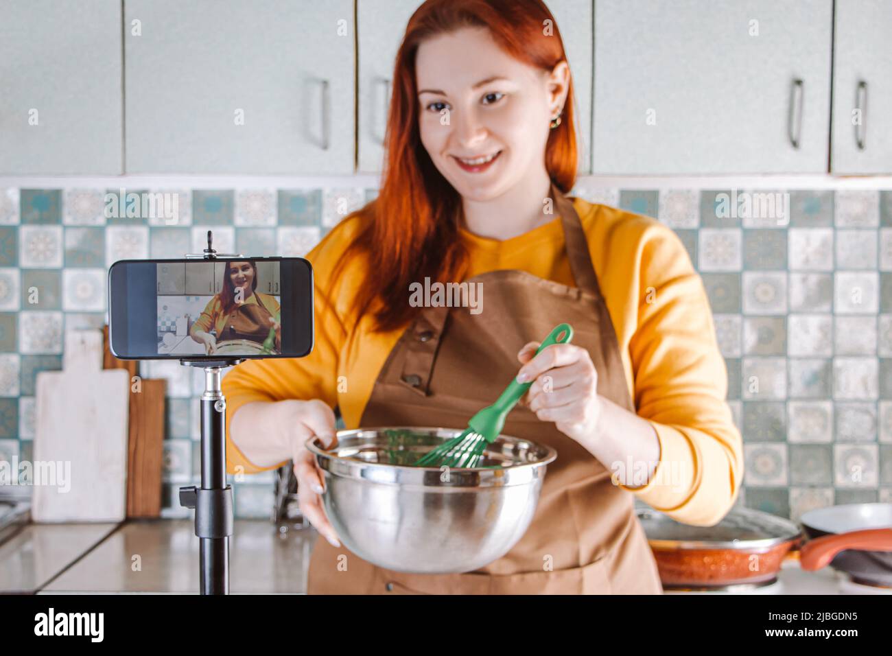 Modern woman blogger is making video for food blog. Young red-haired woman in yellow apron cooking food in home kitchen. Lifestyle, home leisure conce Stock Photo