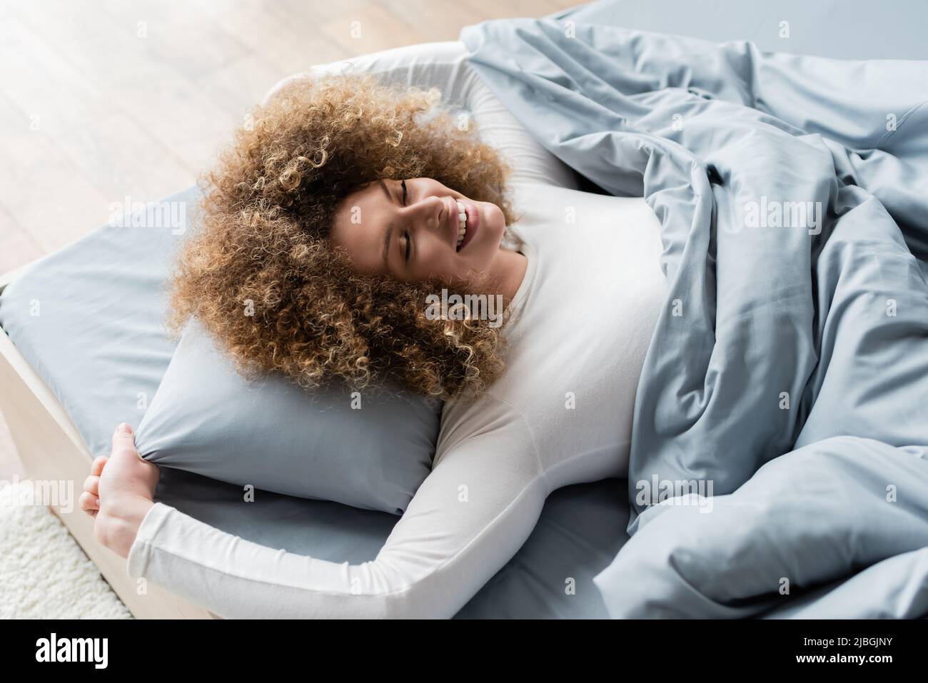 high angle view of joyful woman with wavy hair lying on grey bedding with closed eyes Stock