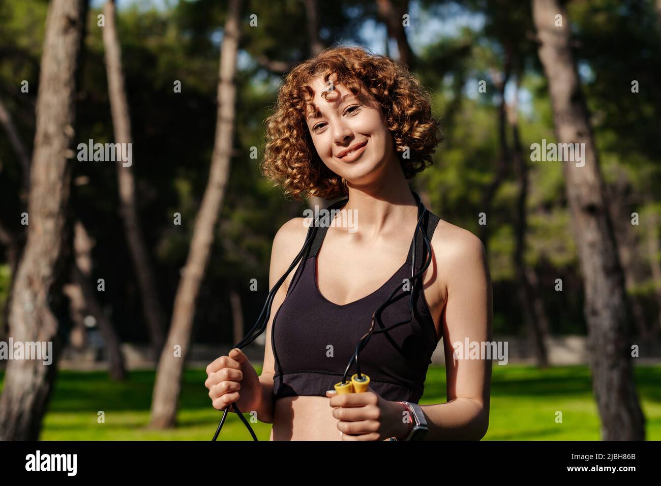 Portrait of young redhead woman wearing black sports bra standing on city  park, outdoors holding a skipping rope on her neck looking at the camera  dur Stock Photo - Alamy