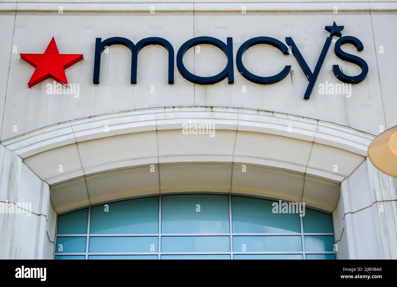 Macy's exterior facade brand and logo signage in thin, black letters with a red star logo on beige stone building above opaque glass windows. Stock Photo