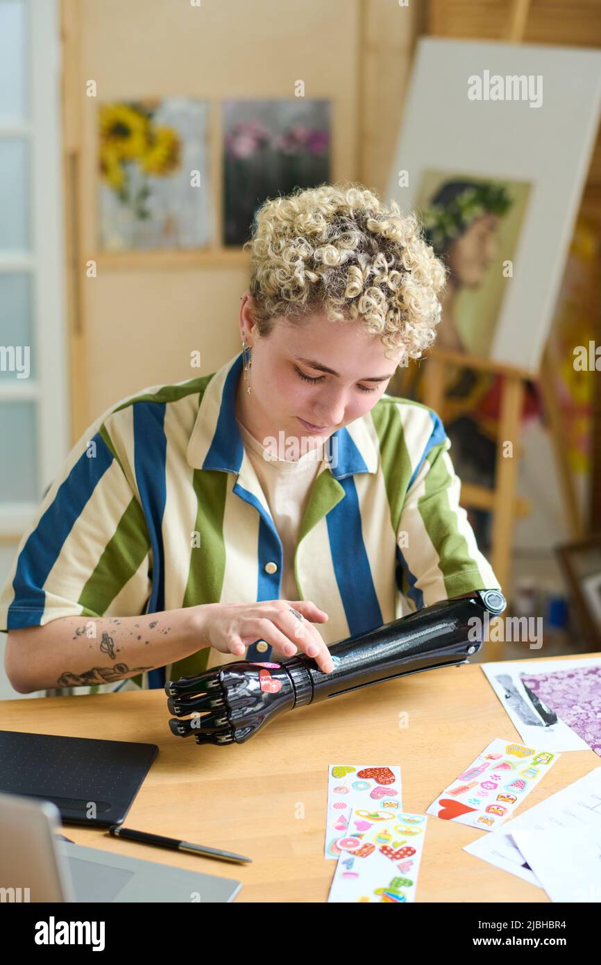 Young woman with disability sticking handmade stickers on her myoelectric arm while sitting by table in living room Stock Photo
