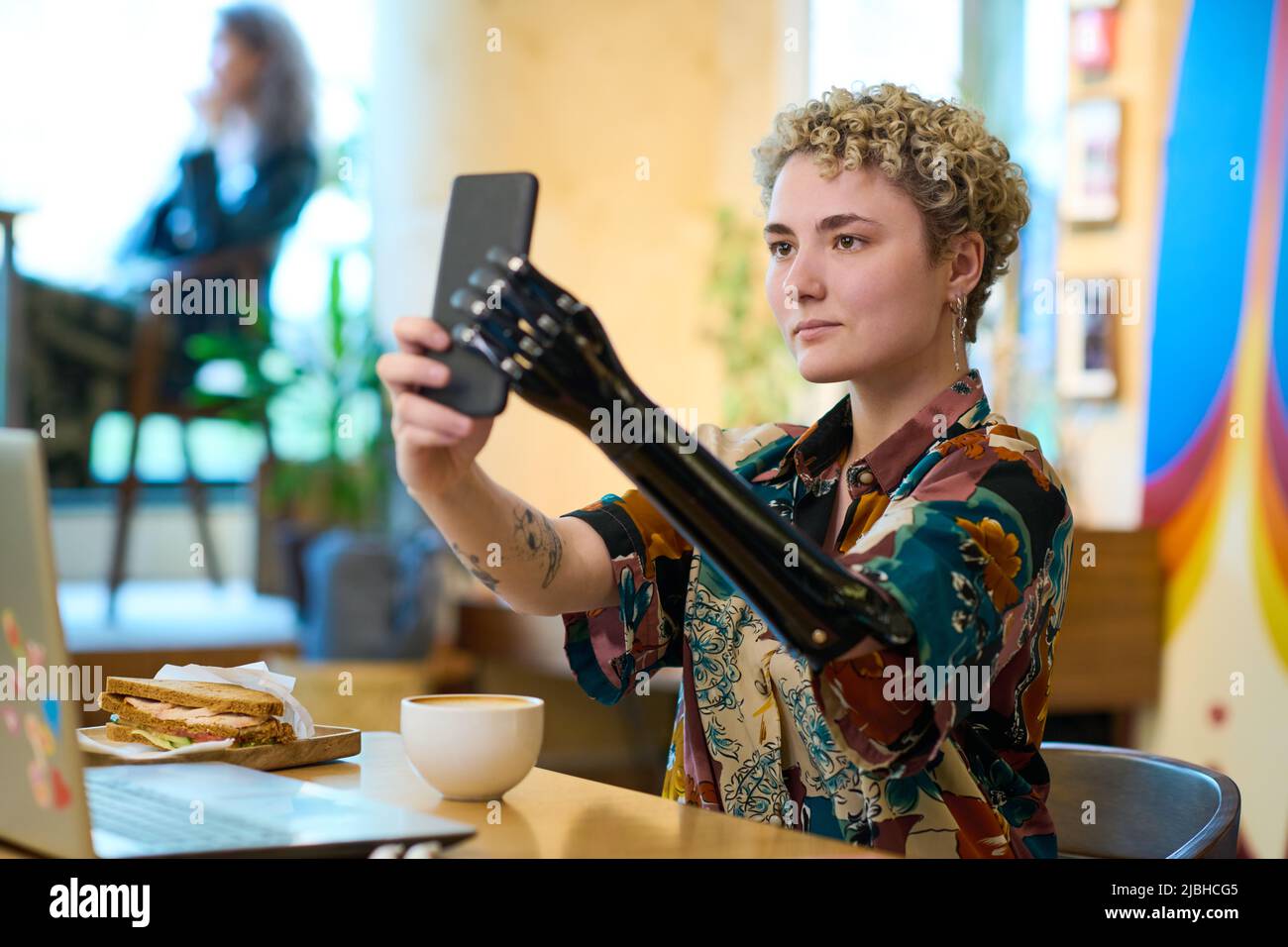 Young woman with robotic arm looking at smartphone camera while making selfie or communicating in video chat during breakfast Stock Photo