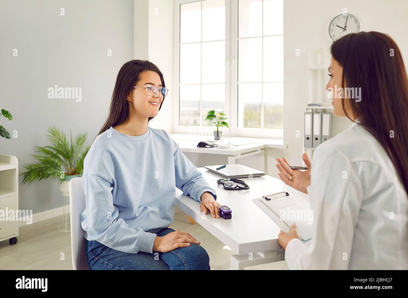 Doctor talks to young female patient who uses fingertip pulse oximeter in medical office. Stock Photo