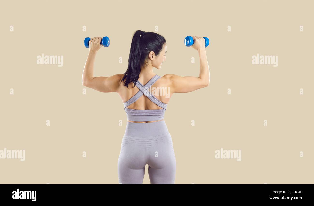 Happy sporty woman lifts small dumbbells while standing with her back to camera on beige background. Stock Photo