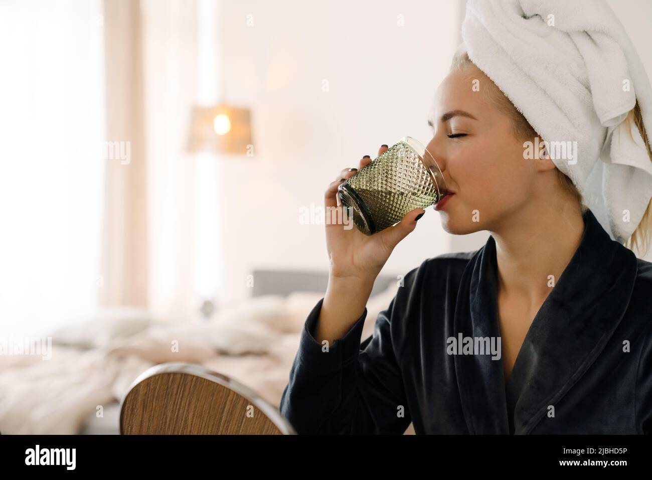 White woman wrapped towel drinking water after shower at home Stock
