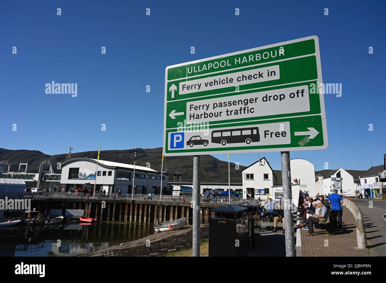 Sign for Ullapool harbour in Scotland, UK, showing arrows to ferry areas. People and ferry buildings in background. Blue sky, sunny day. Stock Photo