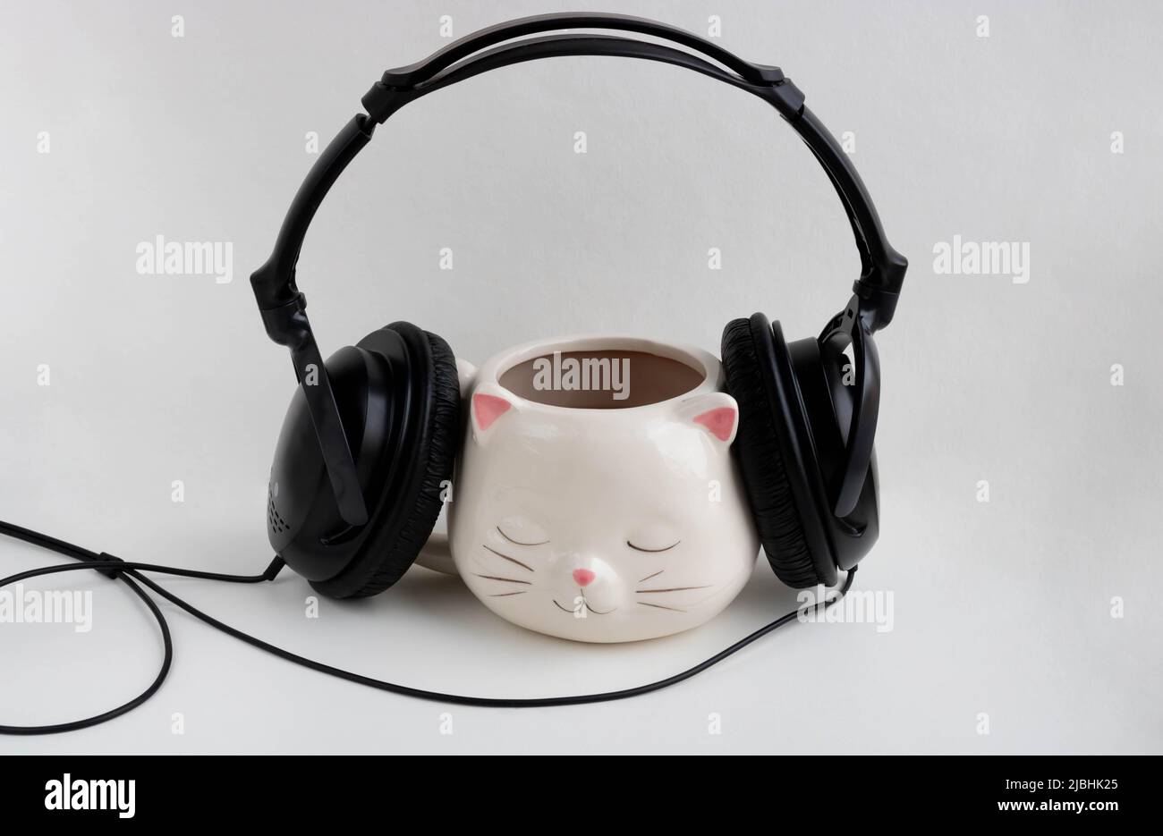 Black headphones are worn on a white coffee mug in the shape of a cat with pink ears, close-up on a white background. Stock Photo