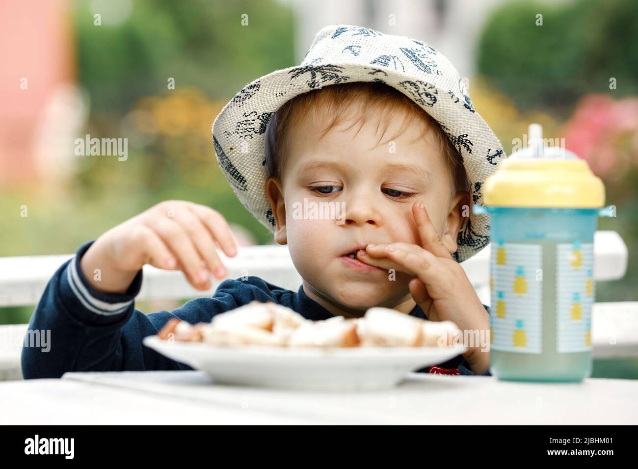 A little boy in a white hat tastes bread with cheese while licking his fingers. A plastic cup with pineapple juice on the table. Stock Photo